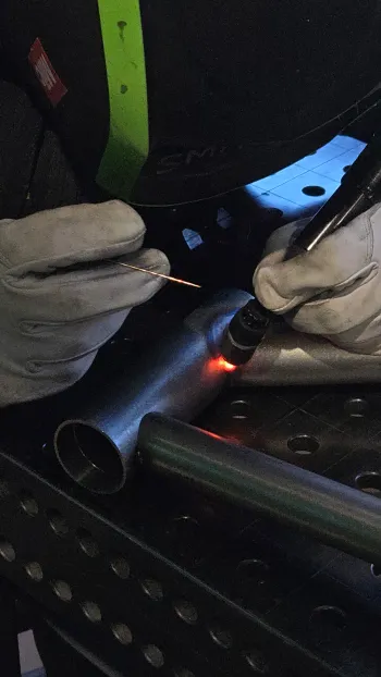 Soudure TIG en cours sur tube de cadre chromoly, mains gantées, torche à l'arc