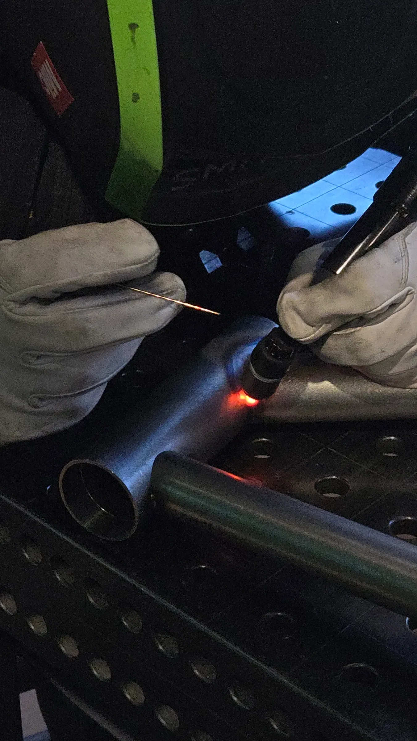 Mains gantées positionnant la torche TIG et la baguette d'apport sur un tube d'acier encore rouge